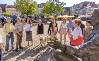 Das Bild zeigt eine Gruppe Menschen, die an einer Stadtführung teilnehmen und am Historischen Marktplatz  in Peine an der Skulptur des Friedrich von Bodenstedt stehen. 